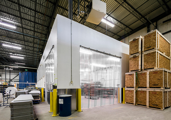 Photo architecturale intérieure dans une industrie, montrant l'accès à une chambre froide, des caisses d'équipement empilées d'un côté et des tables de travail de l'autre