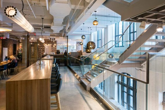 espaces communs d'un bureau du centre ville de Montréal, montrant une longue table en bois, des tabourets, un grand escalier montant à l'étage et des luminaires suspendus