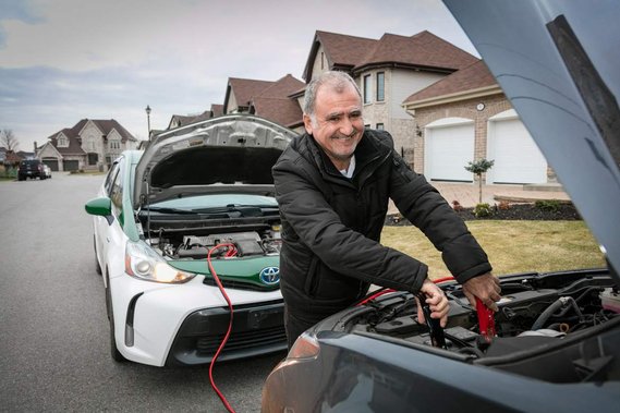 Chauffeur de Taxi COOP offrant le service de survoltage des batteries souriant avec cartier résidentiel à l'arrière plan