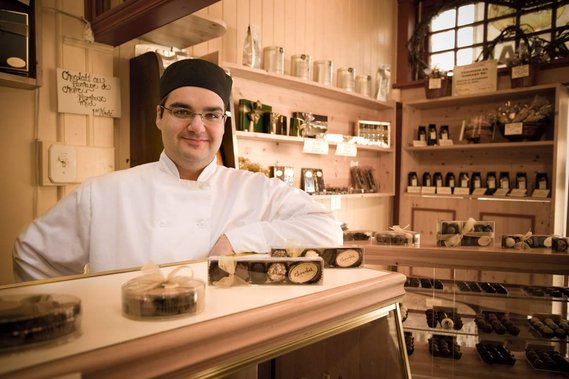 Portrait éditorial d'un pâtissier dans sa boutique, souriant, appuyé sur son comptoir, entouré des produits qu'il a faits 