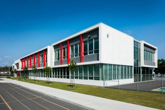 École primaire photographiée en plein soleil pour en montrer la façade et le côté, l'aménagement paysager est soigné, de jeunes arbres ornent la pelouse