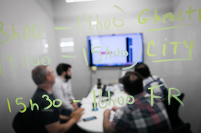 Employés en action ou en pause dans un bureau d'une entreprise  de technologies de l'information, on les aperçoit en réunion derrière une vitrine où sont inscrits des informations au crayon feutre