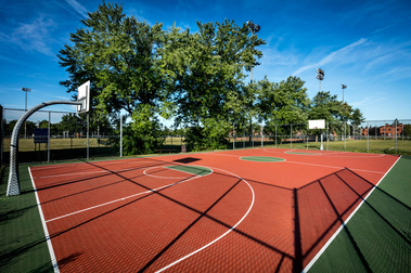 Terrain de basketball extérieur, entouré d'arbres matures et d'une clôture dont la lumière du soleil levant projette l'ombre