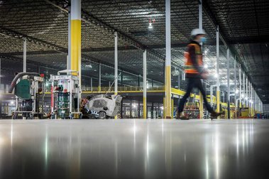 Vue au ras du sol d'un plancher de béton poli, sur un chantier de construction, avec ouvrier qui marche flou et immenses structures d'acier à l'arrière
