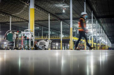 Vue au ras du sol d'un plancher de béton poli, sur un chantier de construction, avec ouvrier qui marche flou et immenses structures d'acier à l'arrière