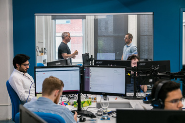Employés en action ou en pause dans un bureau d'une entreprise  de technologies de l'information, disposés devant leurs ordinateurs autour d'une table commune, bureau des patrons qui discutent à l'arrière plan