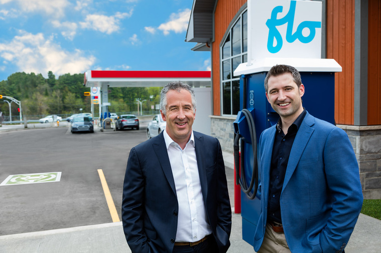 Portrait corporatif de deux dirigeants d'une entreprise de bornes électriques dans une station service, devant une station de recharge flo et des pompes à essence à l'arrière plan