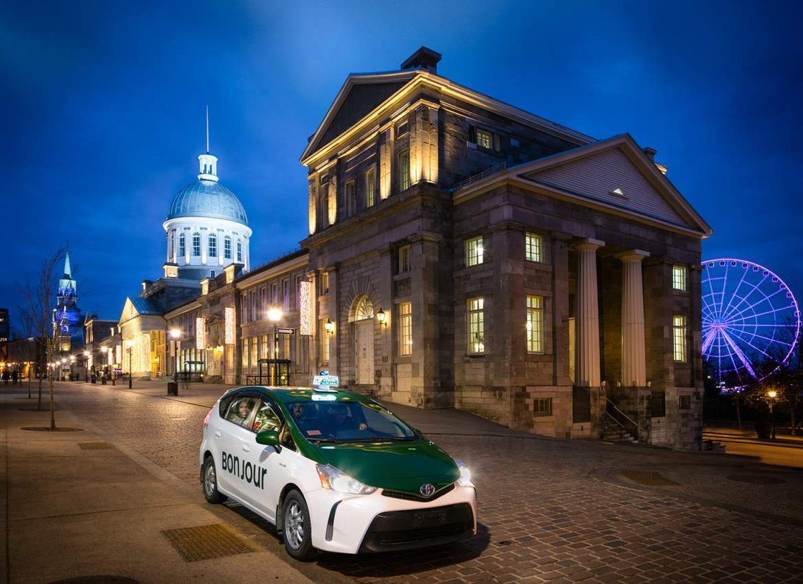 Service de visite guidée pour touristes dans le Vieux-Montréal offert par COOP Taxi, une voiture avec un couple de touristes devant le marché Bonsecours, la grande roue est visible à l'arrière plan, ainsi que le dôme emblématique du bâtiment historique
