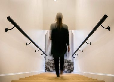 Jeune femme vue de dos, descendant un escalier de bois, avec rampes courantes en bois noire de part et d'autres 