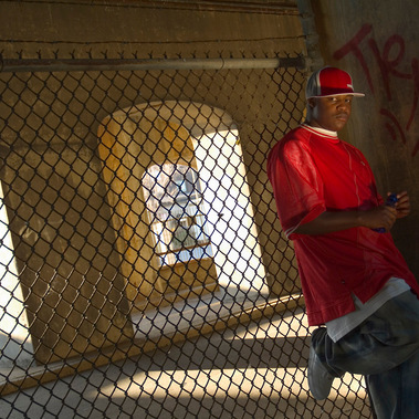 Photo d'un rappeur dans un environnement urbain, appuyé sur une clôture métallique, il fixe l'objectif, casquette sur la tête