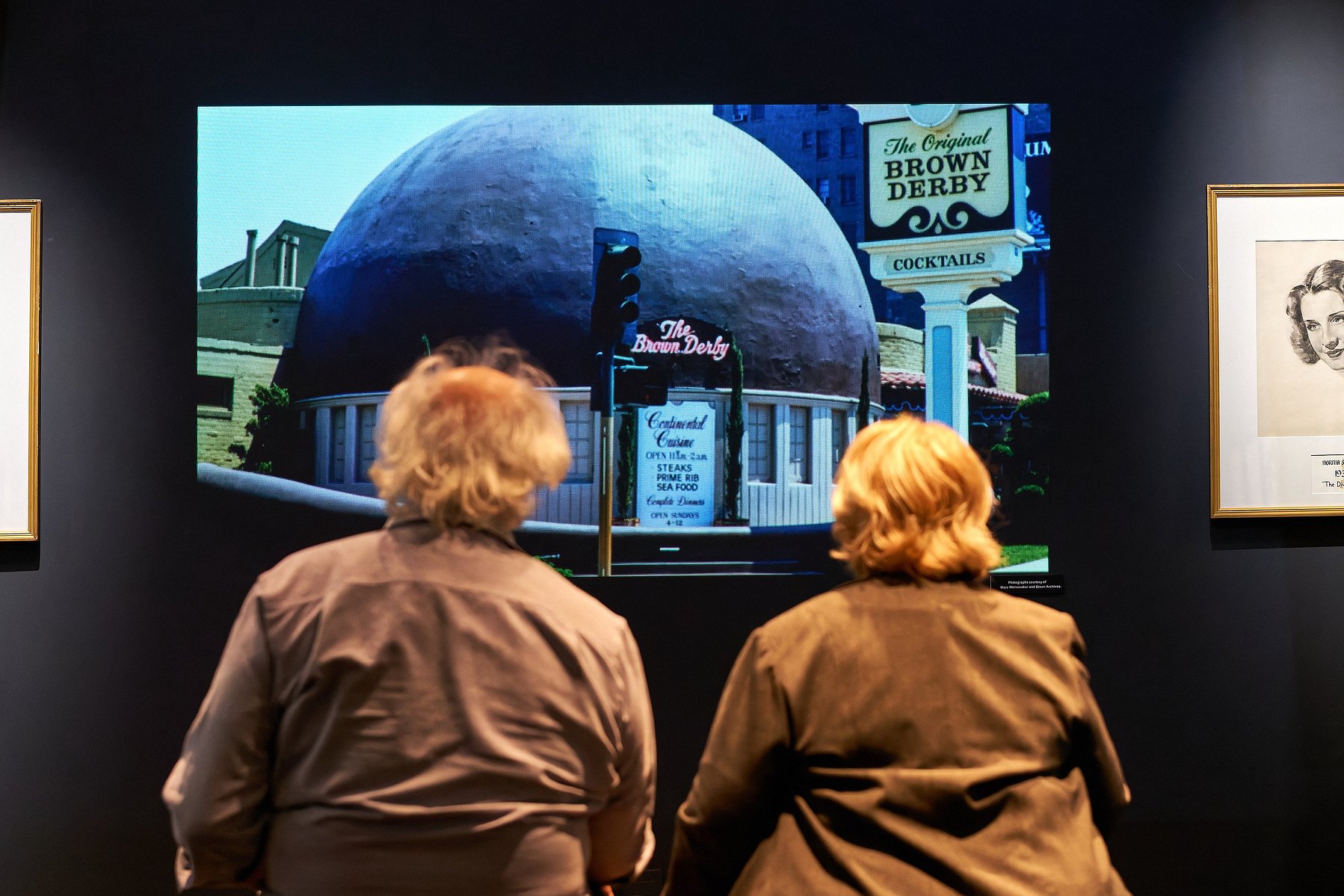 Two people sit and watch a video about the Brown Derby  at "Painted for the Pictures" at Muzeo in Anaheim, CA