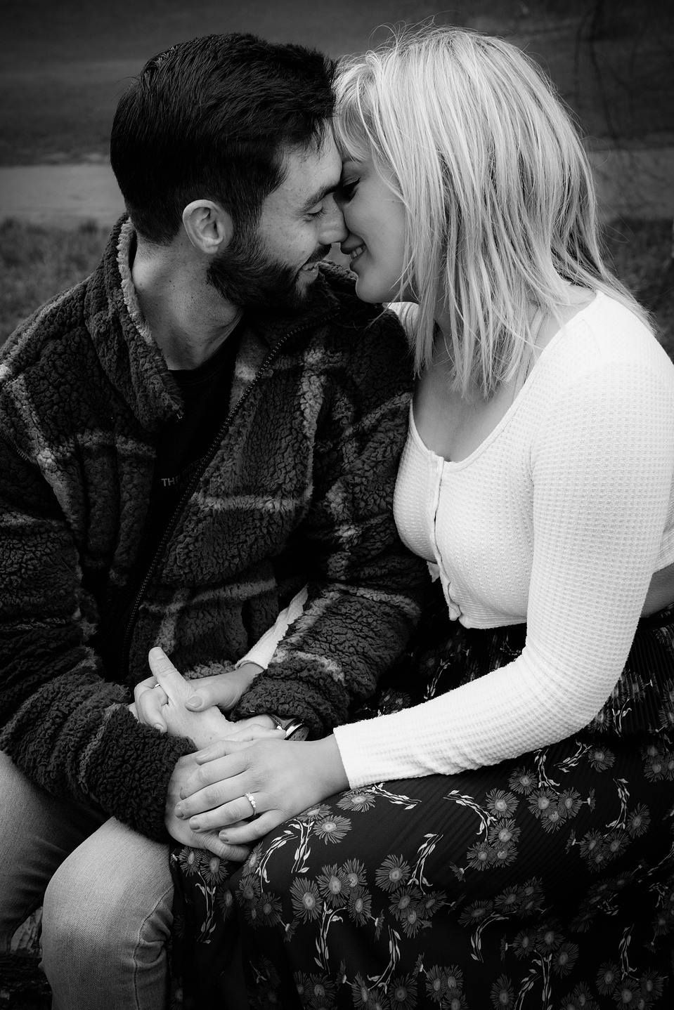 A couple sitting down, about to kiss and smiling. Hands are in laps and she has a new engagement ring on.