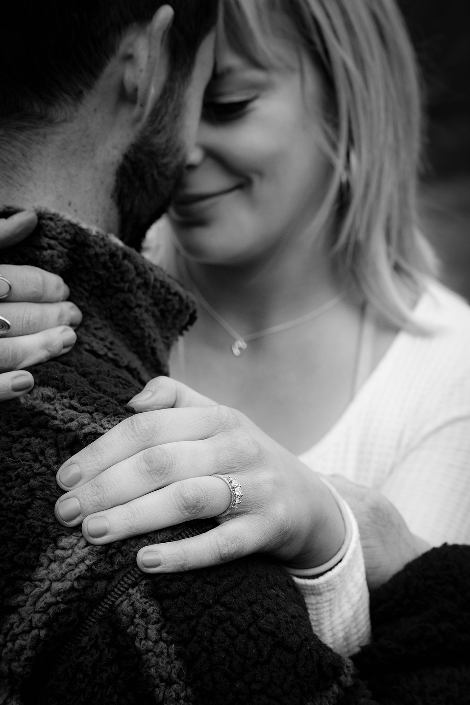 A couple embracing and her hand on his neck and shoulder, smiling at each other and showing off her new engagement ring