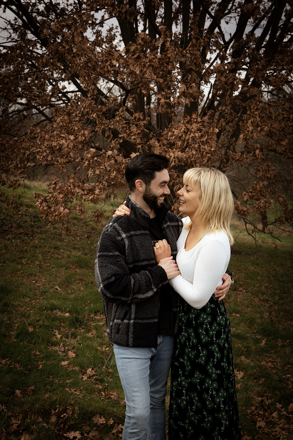 A newly engaged couple embracing each other, looking at each other and smiling. In a field in Greenwich, London. 