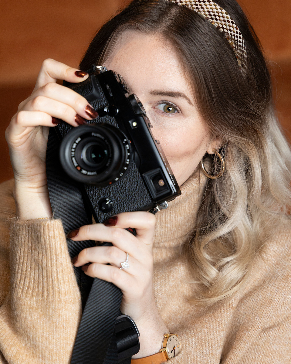 Woman holding a camera over her face but one eye peeking through wearing a beige jumper and headband