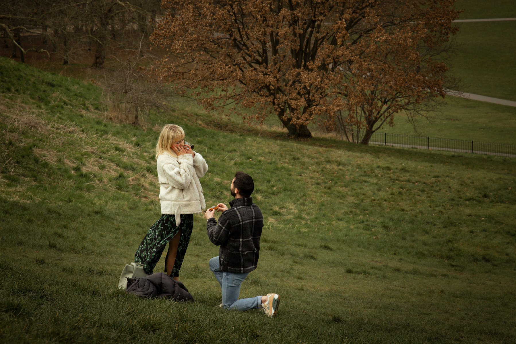 Man on one knee proposing to a lady. She is so shocked and excited. In Greenwich, London in the middle of Greenwich park. 