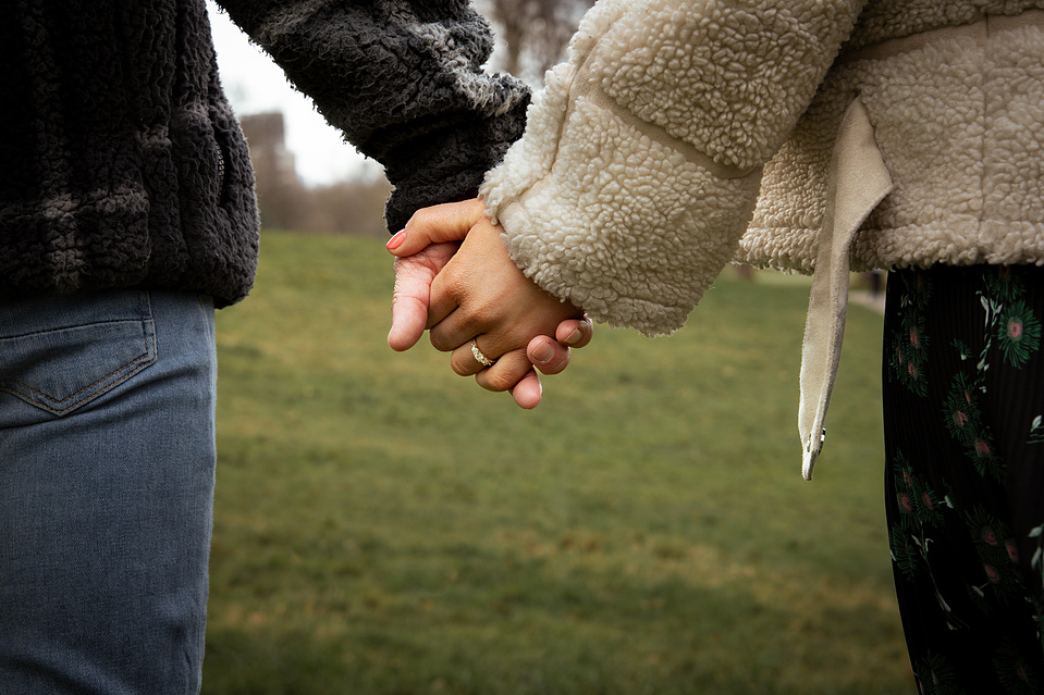 A couple holding hands in a field, she has an engagement ring on her finger. 