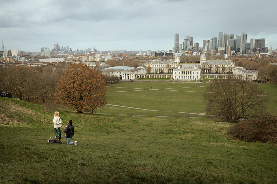 A man on one knee proposing to a lady in the middle of Greenwich park, London. The city in seen in the background.