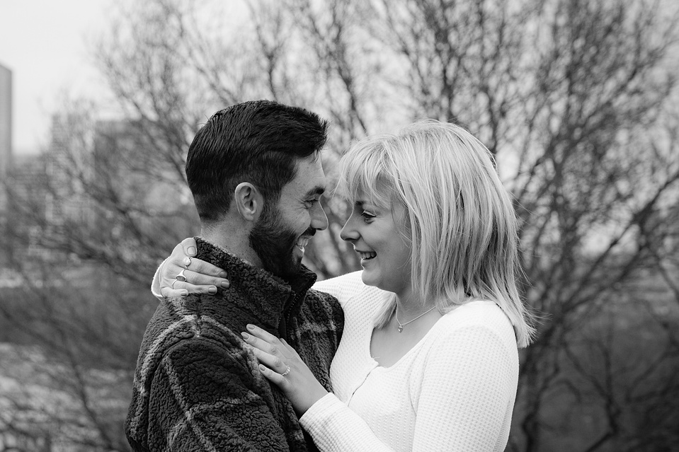 A couple smiling at each other, her hands around his neck, their faces close after thay have just got engaged. 