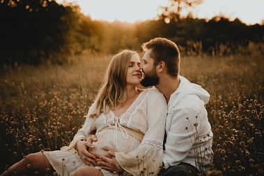 Bei einem Babybauch Fotoshooting in Berlin sitzt ein Paar auf einer Wiese und küsst sich im Sonnenuntergang. 