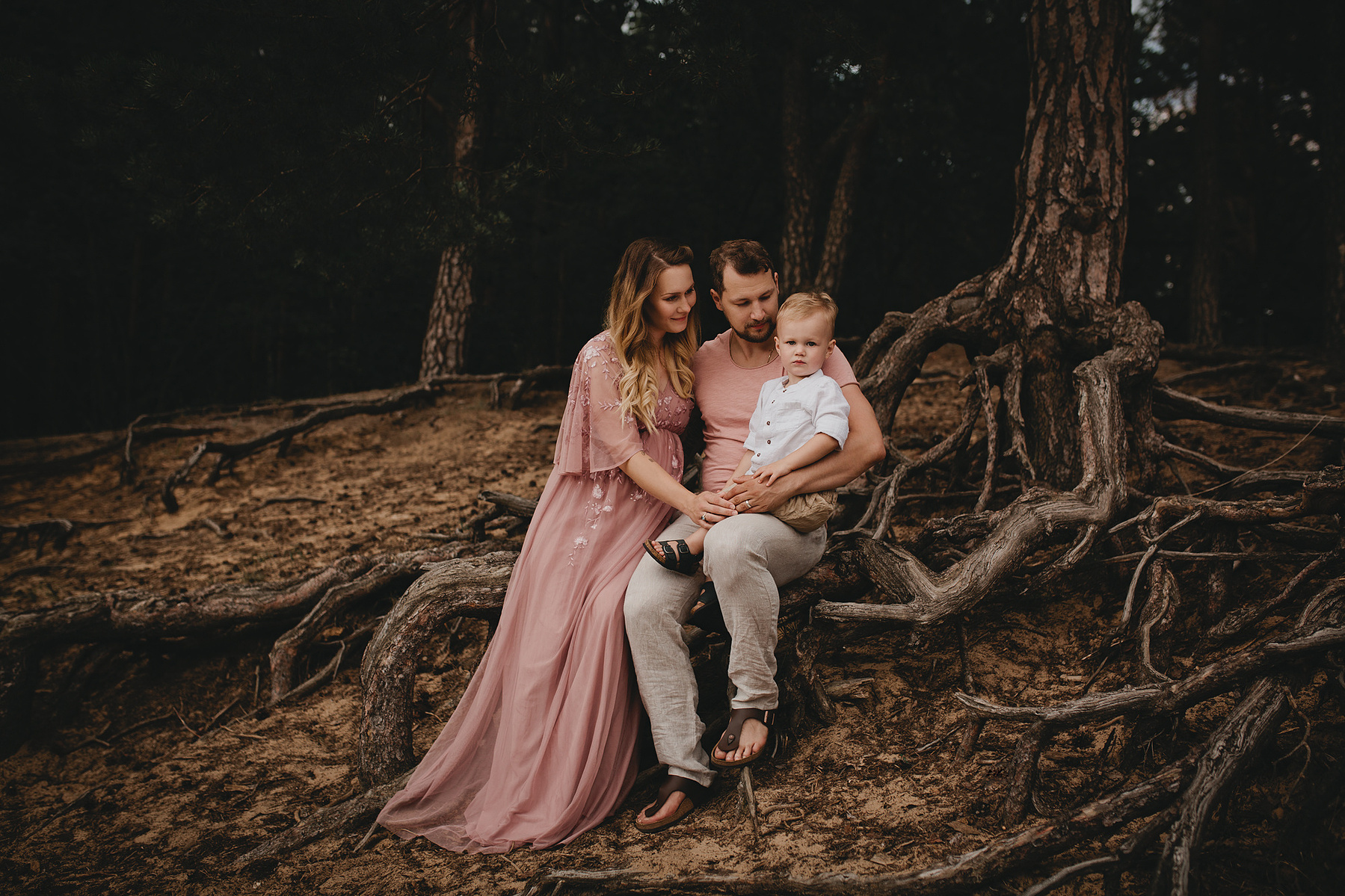 Familie mit Outfits in rosa und beige sitzt beim Fotoshooting auf Wurzeln im Wald in Brandenburg.