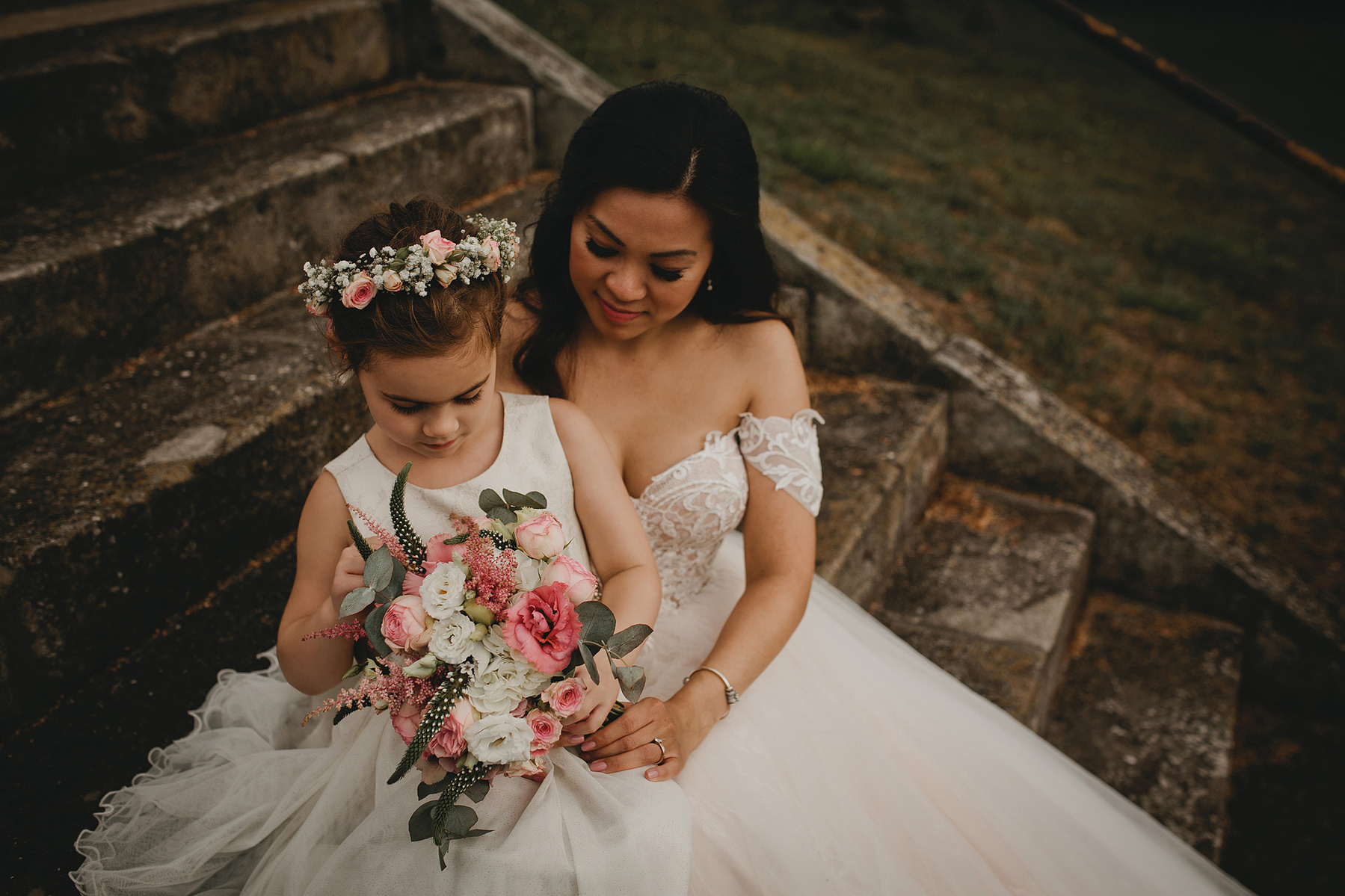 Eine Braut im weißen Hochzeitskleid sitzt mit ihrer Tochter auf einer Treppe. Das Kind betrachtet neugierig den Brautstrauß.