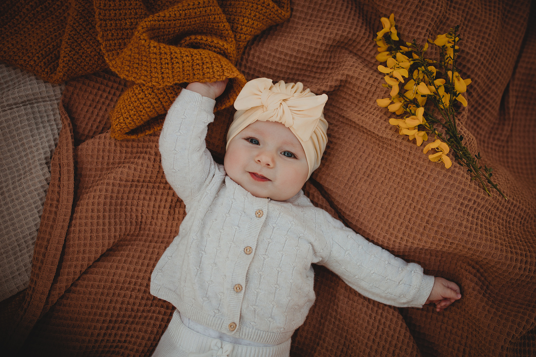 Ein Baby im cremeweißen Outfit liegt für Kinderfotos im Bohostil auf einer mit Blumen dekorierten Decke. 