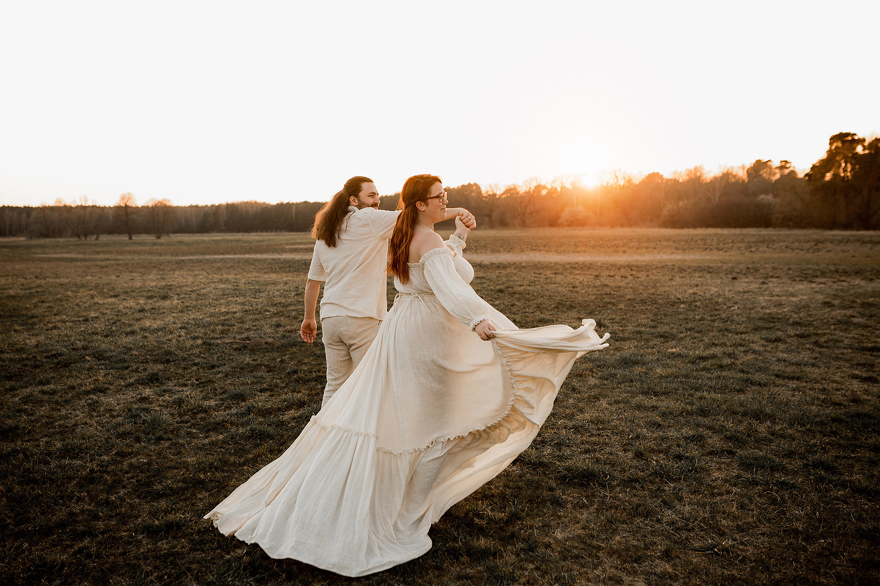 Für natürliche Babybauchfotos in Berlin läuft ein Paar im Abendlicht über eine Wiese. Das Babybauchkleid im Bohostil weht im Wind. 