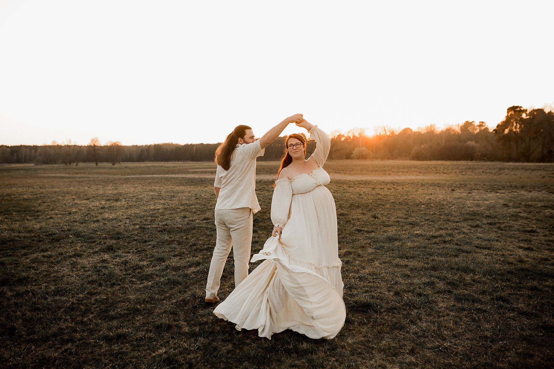 Beim Babybauch Fotoshooting in Berlin tanzt ein Mann mit seiner schwangeren Frau im Sonnenuntergang auf einem Feld. 