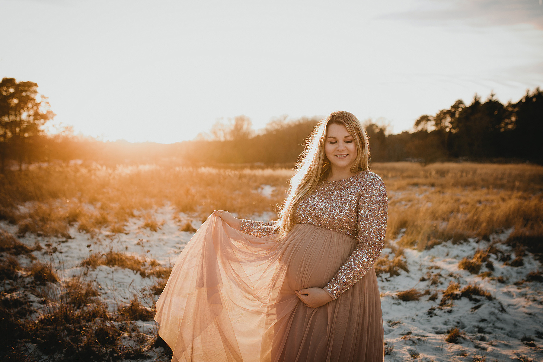 Beim Schwangerschafts-Fotoshooting im Winter steht eine Frau mit Babybauch auf einer verschneiten Wiese und lässt ihr Kleid im Wind wehen.