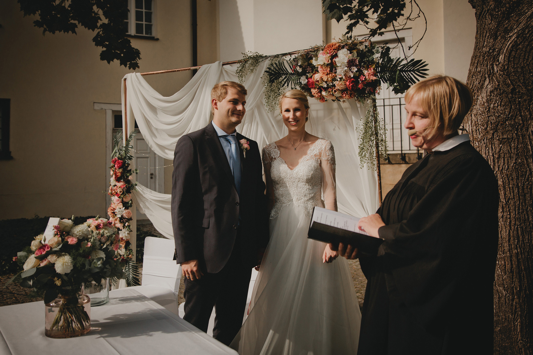 Das Brautpaar steht beim Jawort vor dem mit Blumen und Chiffon verzierten Traubogen aus Kupferrohren. 