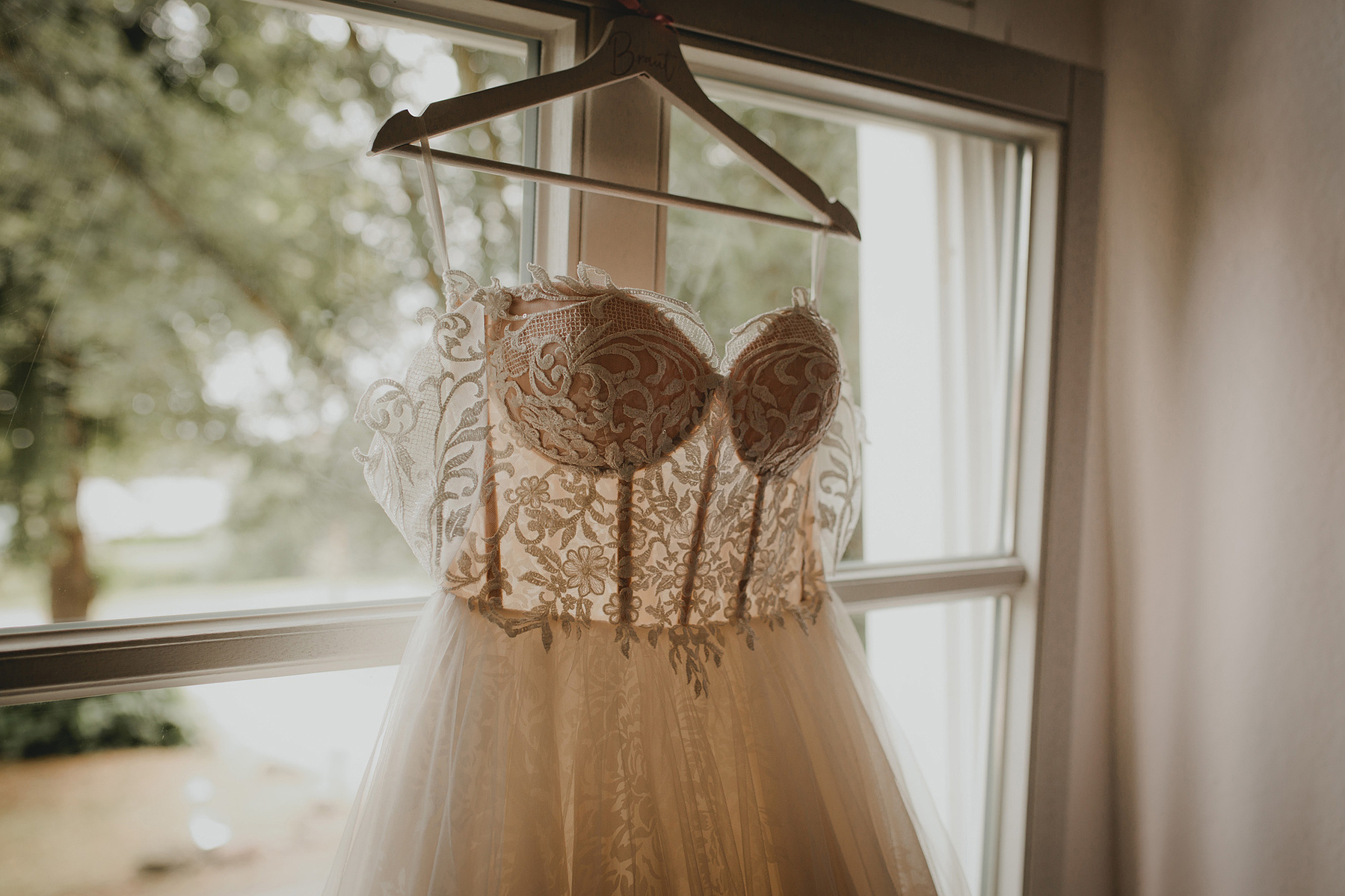 Ein cremefarbenes Brautkleid mit Corsagentop und Spitze hängt vor einem Fenster.