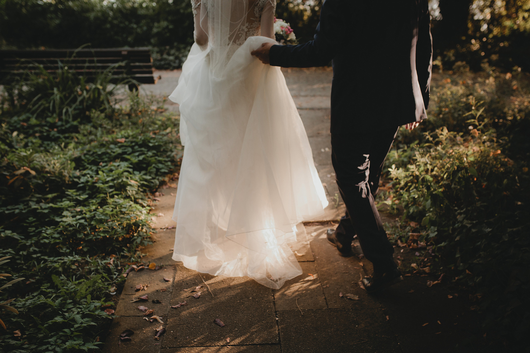 Der Bräutigam hebt die Schleppe des Hochzeitskleides an während die Braut durch den Park läuft.