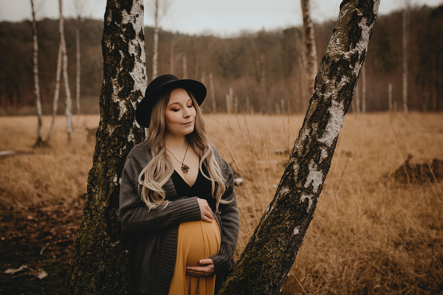 Eine Schwangere lehnt beim Fotoshooting in Berlin an einem Baum und blickt liebevoll auf ihren Babybauch.
