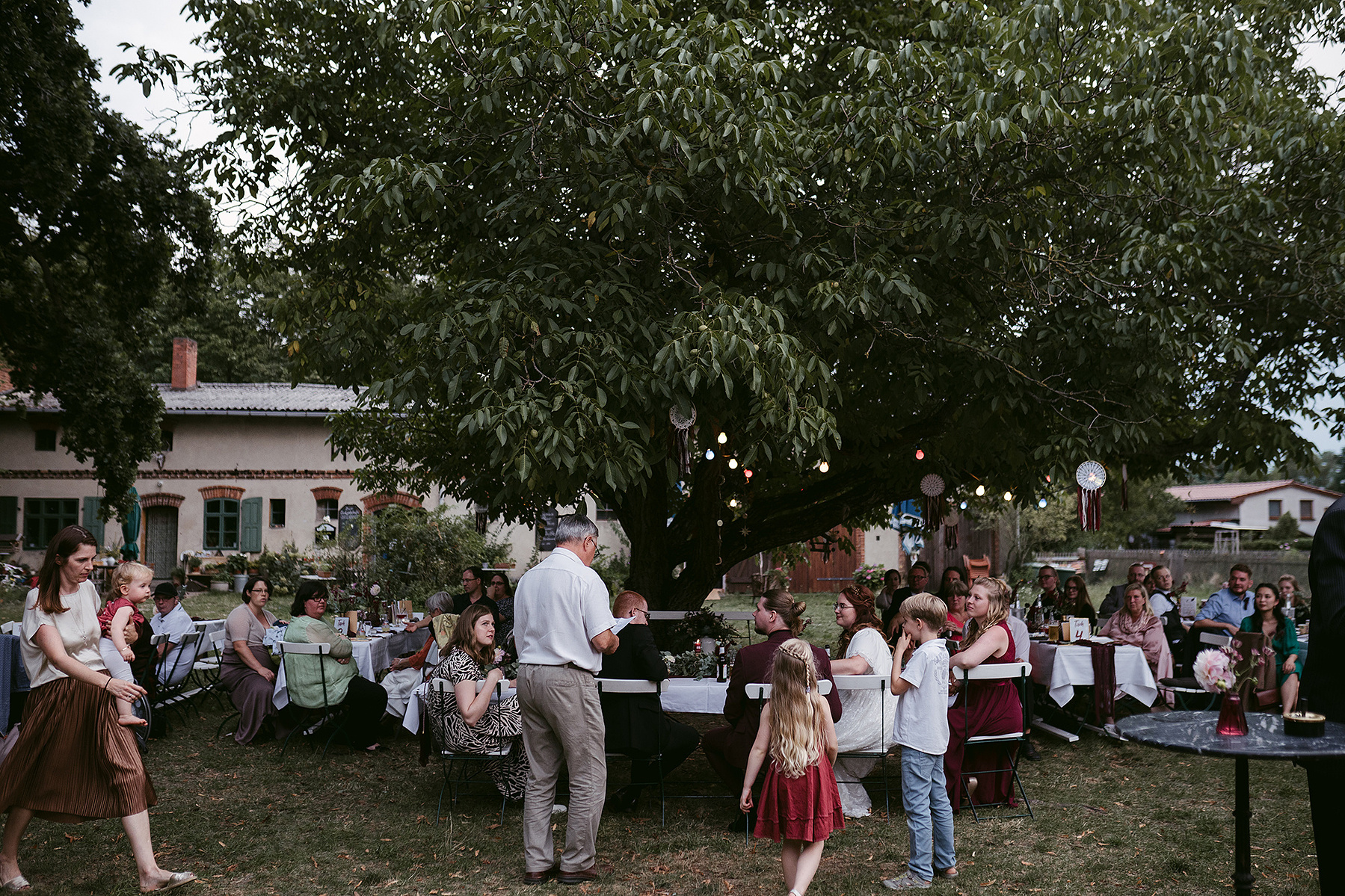 Sommerhochzeit im Garten der Alten Försterei Briescht. Am Abend sitzen die Hochzeitsgäste im Hof unter einem großen Baum mit Lichterketten.