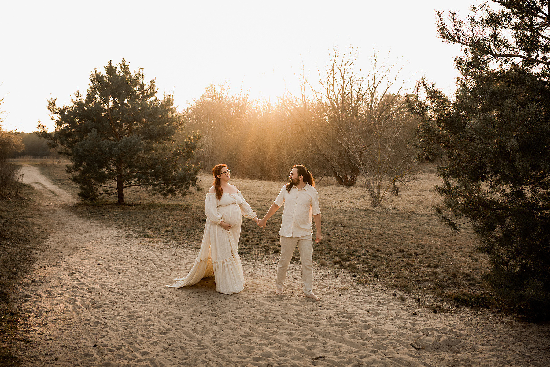 Bei einem Babybauchshooting in der Natur läuft ein Pärchen in boho Outfits barfuß durch den Sand. 