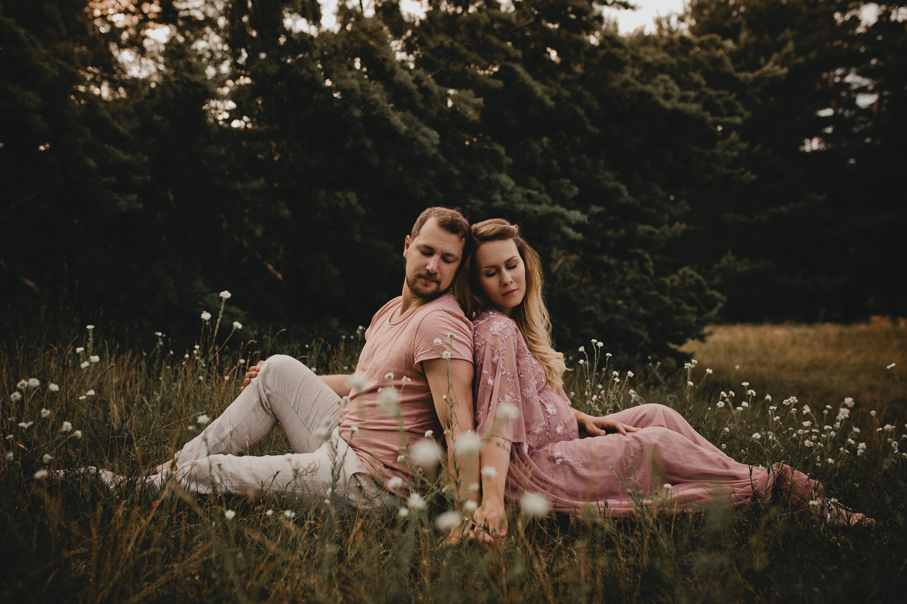 Jana und Chris sitzen beim Paarshooting mit Babybauch in Berlin Rücken an Rücken auf einer blühenden Wiese.