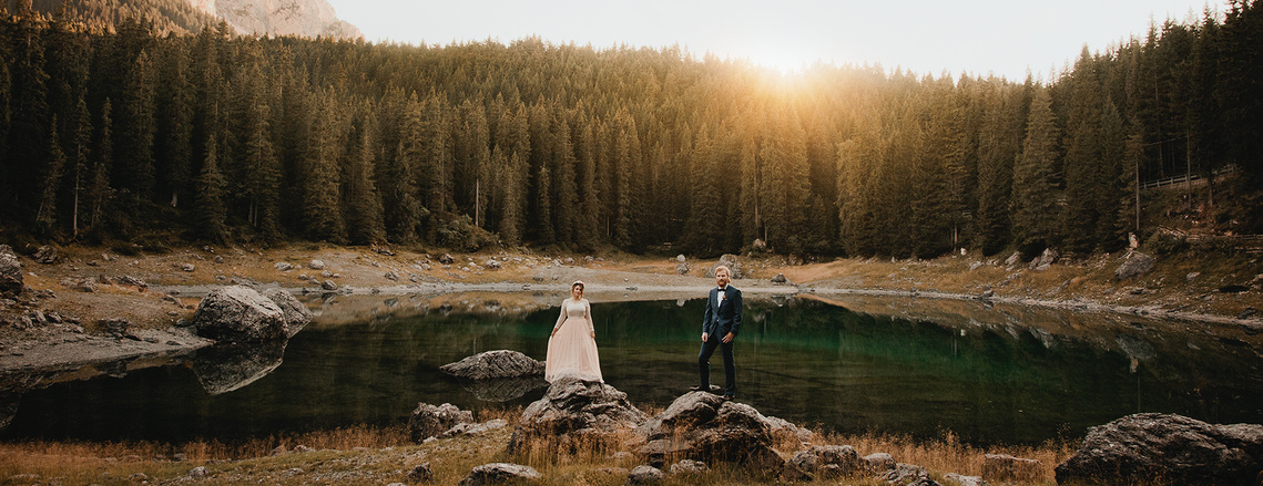 Elopement in den Alpen. Hochzeitspaar steht im Sonnenuntergang auf Felsen vor einem Bergsee.