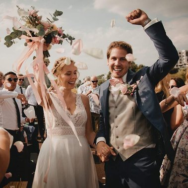 Jubelndes Brautpaar beim Auszug nach der Hochzeit auf einem Boot in Berlin. Die Braut hält ihren Brautstrauß hoch, während die Hochzeitsgäste Blütenblätter werfen.