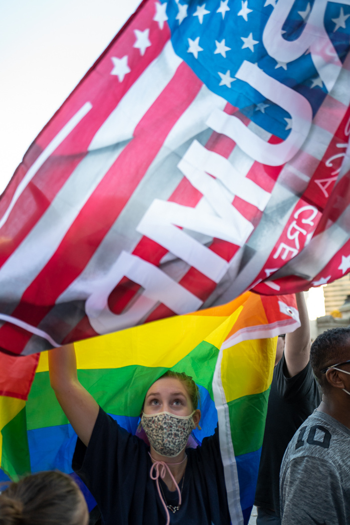 A girl holding LGBTQ pride flag, Trump flag flies above it