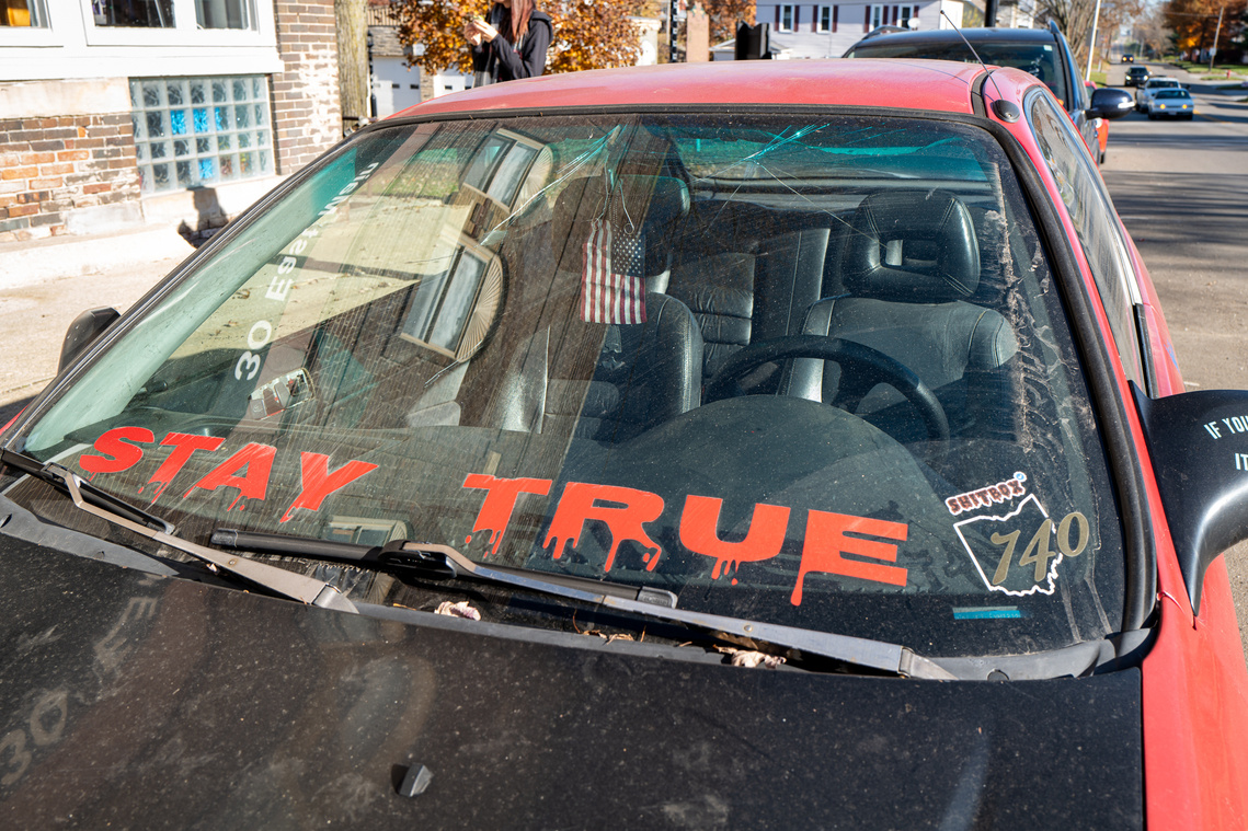 A “Stay true” sign on a windshield, an American flag hanging in the red car parked on a street in a small town, before the 2020 United States presidential election
