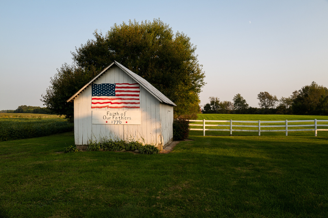  A “Faith of our fathers” sign and an American flag painted on a white shed illuminated by evening light, surrounded by idyllic countryside, before the 2020 United States presidential election