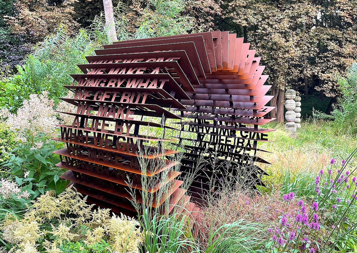 Mantra Machine Corten sculpture nestled in a beautiful collectors sculpture garden in Yorkshire during summer, surrounded by  flowers, trees and overgrown grass. 
