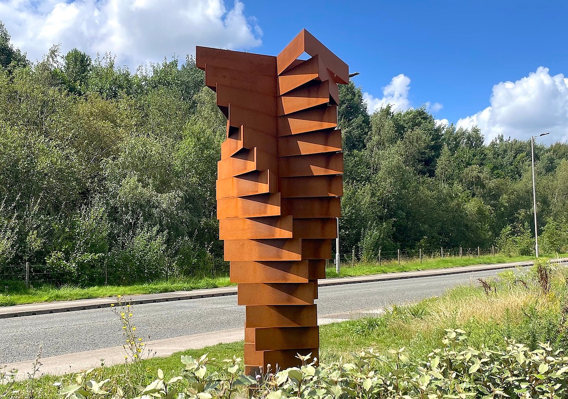 Tall twisting abstract sculpture made from Corten Steel. installed next to a road surrounded by grass, trees and bushes.