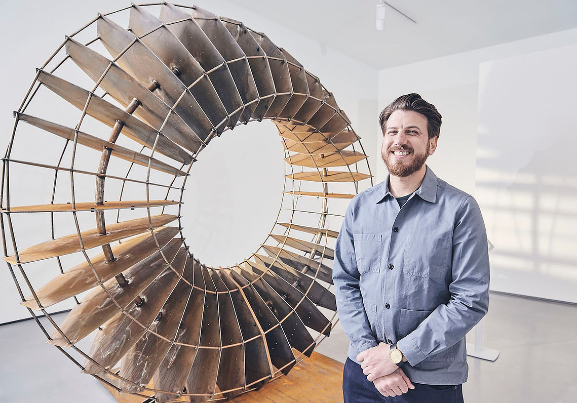 Sapien stands smiling in front of his Only Breath sculpture.  The large Corten sculpture, looks like an oversized machine part. It is circular shaped with a a spiring  form and lots of gaps between the panel blades.