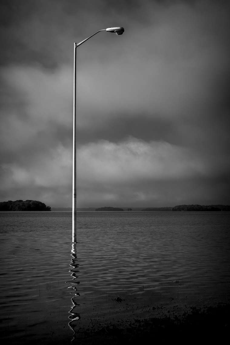 A solitary streetlamp rises from floodwaters under a moody sky, its reflection rippling through the still surface—an eerie yet striking scene of nature reclaiming space.
