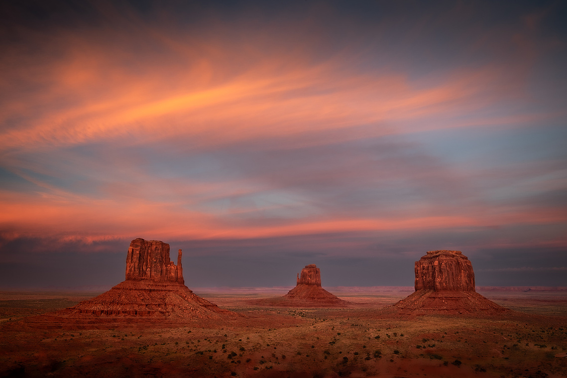 The Mittens and Merrick Butte glow beneath vibrant streaks of pink and orange clouds at sunset in Monument Valley, Utah, standing tall over the vast desert landscape in iconic, timeless beauty.