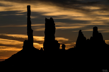Silhouetted rock spires, known as totems, of Monument Valley, Utah, rise against a dramatic sunrise sky, their dark forms etched sharply against the warm golden and amber hues of dawn.