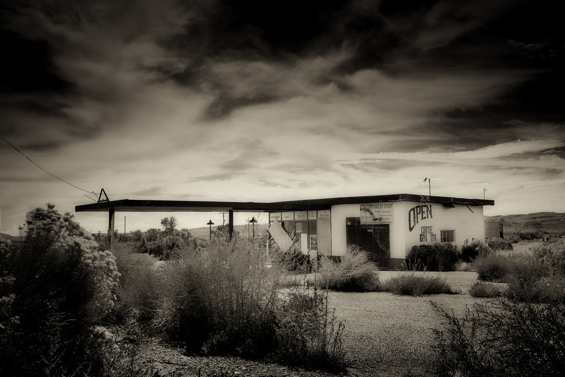 An abandoned gas station in the Utah desert stands weathered and forgotten, its “OPEN—COME ON IN” sign fading into irony. Overgrown brush and cracked pavement encroach on the building, hinting at a bygone era of roadside stops and long stretches of isolat