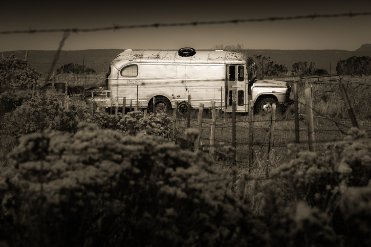 An abandoned bus rests in the desert scrub near Monument Valley, Utah, its weathered frame and peeling paint echoing a bygone era, surrounded by barbed wire and sagebrush under the vast, open sky of the high desert.